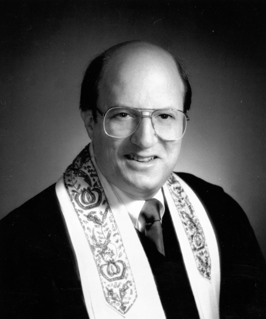 Black and white portrait of a man wearing glasses and a formal robe with a decorative stole, smiling at the camera.