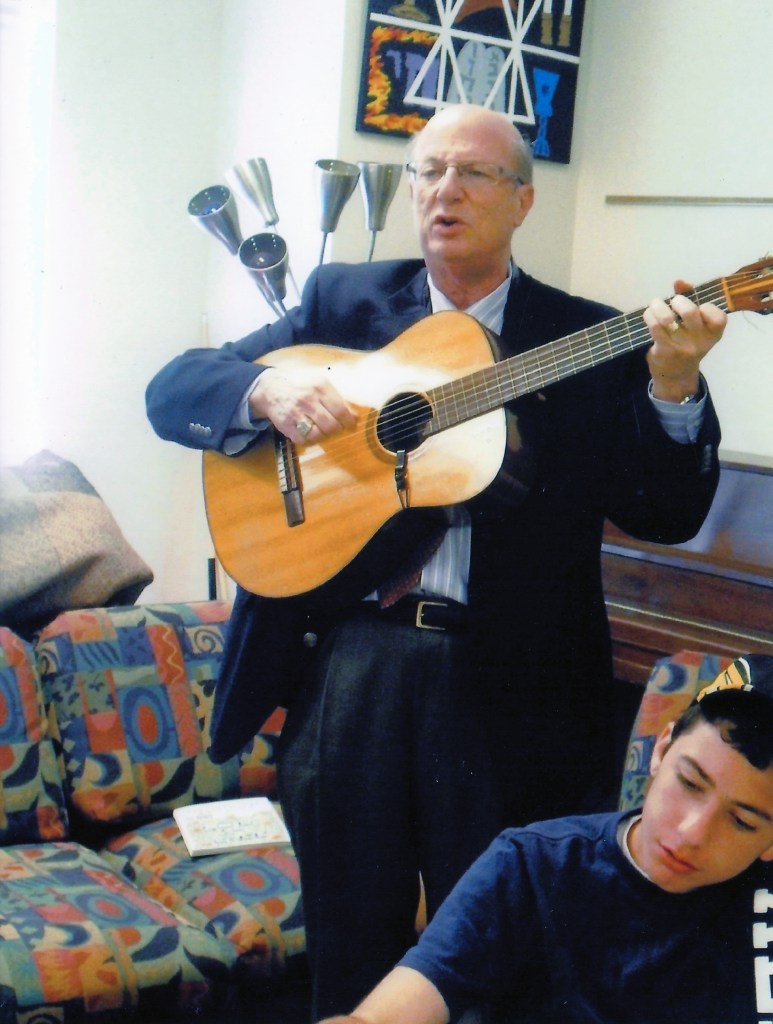 A man in a suit playing guitar and singing while standing in a living room, with a young man sitting nearby, looking engaged. The background features colorful furniture and art.