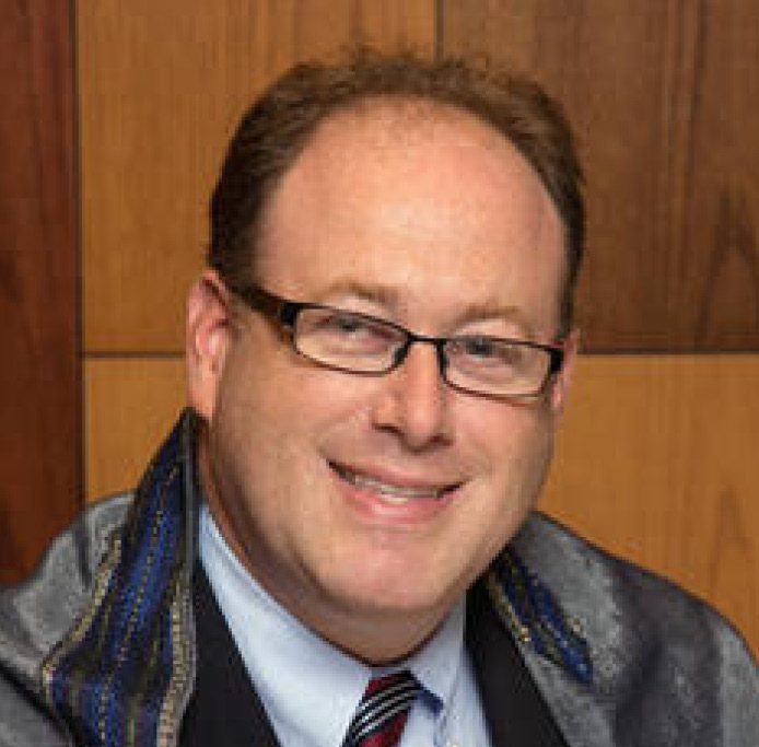 A man with glasses smiling, wearing a formal shirt and a shawl, against a wooden background.