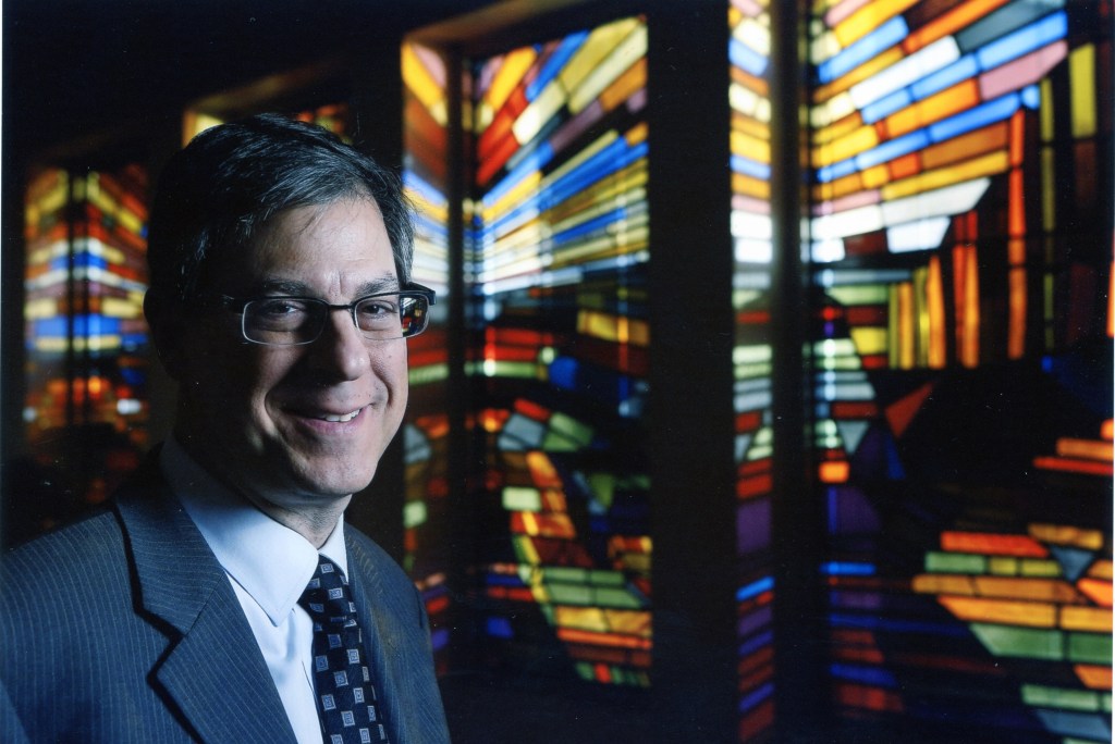 A man in a suit stands smiling in front of vibrant stained glass windows, showcasing a variety of colors and patterns.