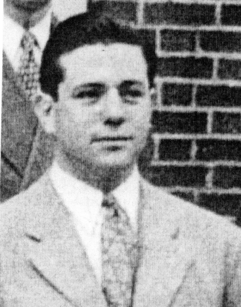 Black and white portrait of a man in a suit with a tie, standing in front of a brick wall.